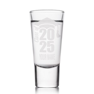 May include: Clear shot glass with etched design. The design includes a graduation cap, the text "Class of 2025", and space for a name. The glass is cylindrical with a thick base and is filled with a clear liquid.