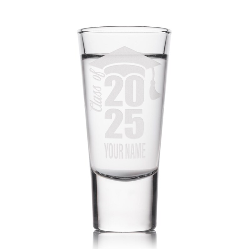 May include: Clear shot glass with etched design. The design includes a graduation cap, the text "Class of 2025", and space for a name. The glass is cylindrical with a thick base and is filled with a clear liquid.