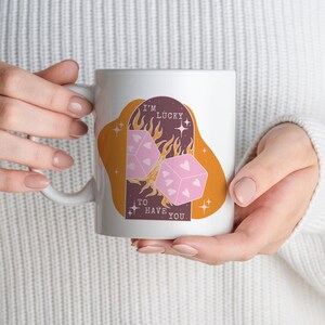 May include: White ceramic mug with a graphic of two pink dice with hearts on them, a flame, and the text "I'm lucky to have you".