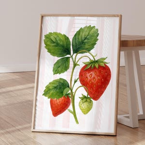 May include: Watercolor print of a strawberry plant with green leaves and three strawberries in varying stages of ripeness. Two are red, and one is green. The background has pink and white vertical stripes, and the print is in a wooden frame.