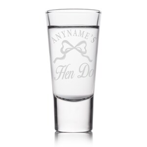 May include: A clear shot glass with etched text that reads "ANYNAME'S Hen Do" with a bow graphic. The glass is cylindrical with a thick base and is designed for a single shot of liquor.