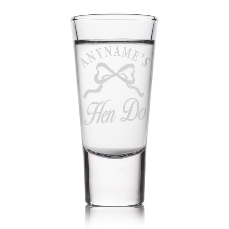 May include: A clear shot glass with etched text that reads "ANYNAME'S Hen Do" with a bow graphic. The glass is cylindrical with a thick base and is designed for a single shot of liquor.