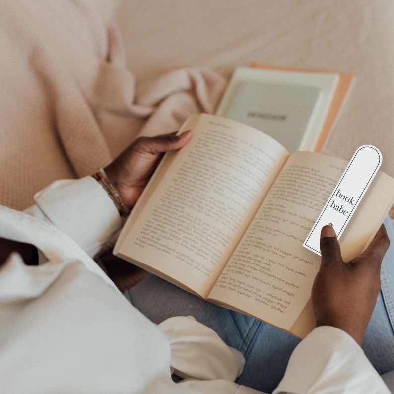May include: A person reading a book with a white bookmark that says "book babe".