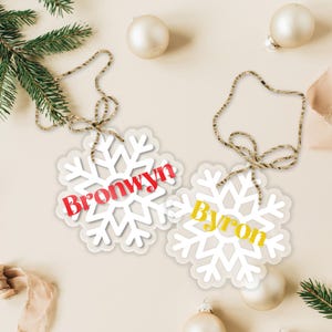 Op de afbeelding: Twee witte sneeuwvlokversieringen met touwtjes om op te hangen. De sneeuwvlok links heeft de naam "Bronwyn" in rode en witte letters. De sneeuwvlok rechts heeft de naam "Byron" in gele letters.