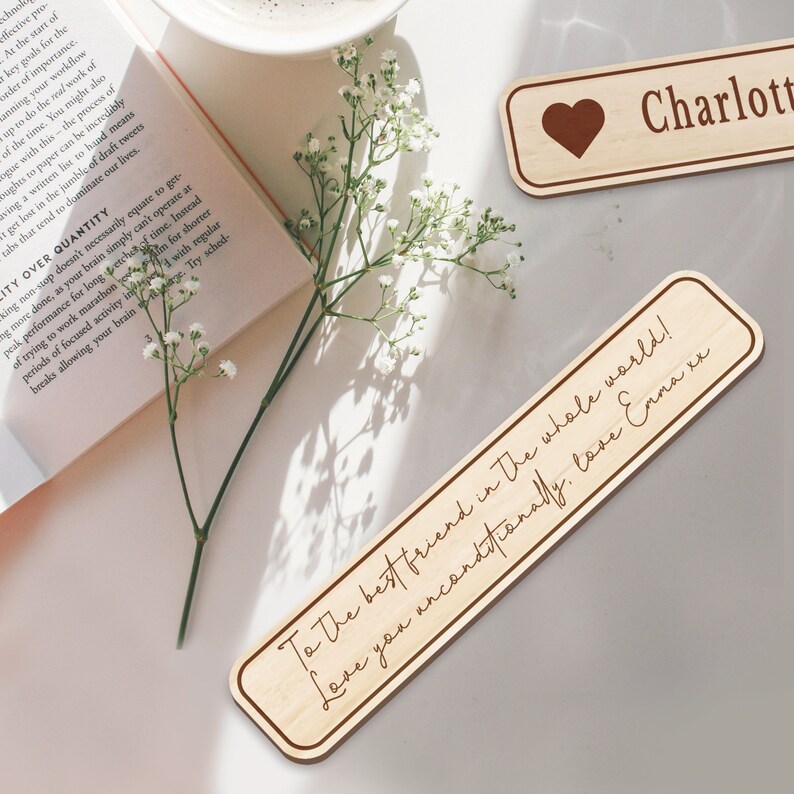 Op de afbeelding: Twee houten bladwijzers met gepersonaliseerde berichten. De eerste bladwijzer is lichtbruin met een hart en de tekst "Charlotte". De tweede bladwijzer is lichtbruin met de tekst "To the best friend in the whole world! Love you unconditionally. Love Emma xx".