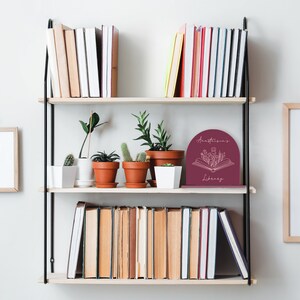 May include: A black metal bookshelf with two shelves. The top shelf has a collection of books and potted plants. The bottom shelf has a collection of books and a burgundy bookend with the text "Anastasia's Library" and a floral design.