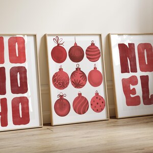 Op de afbeelding: Drie ingelijste prints met een kerstthema. De middelste print toont negen rode kerstballen met verschillende ontwerpen. De andere twee prints tonen de woorden "HO HO HO" en "NOEL" in rode blokletters. De frames zijn licht van kleur.