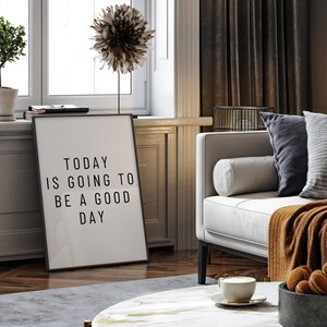 K&ouml;nnte beinhalten: Ein schwarz-wei&szlig;er gerahmter Druck mit dem Text "TODAY IS GOING TO BE A GOOD DAY" lehnt an einer Wand. Das Bild zeigt ein modernes Wohnzimmer mit Sofa, Kissen und einem Couchtisch mit einer Kaffeetasse und Keksen.