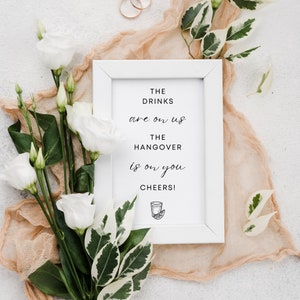 May include: White framed sign with the text "THE DRINKS are on US THE HANGOVER is on YOU CHEERS!" with a small illustration of a margarita glass. The sign is surrounded by white flowers and green leaves.