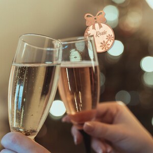 May include: Two champagne flutes filled with sparkling wine, being clinked together in a toast. A wooden Christmas ornament with the name "Rosie" is visible in the background.