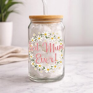 Puede incluir: Un tarro de cristal transparente con tapa de bamb&uacute; y pajita de cristal. El tarro est&aacute; decorado con una corona de margaritas y el texto "Best Mum Ever!" en rosa. El tarro est&aacute; relleno de papel de seda blanco.