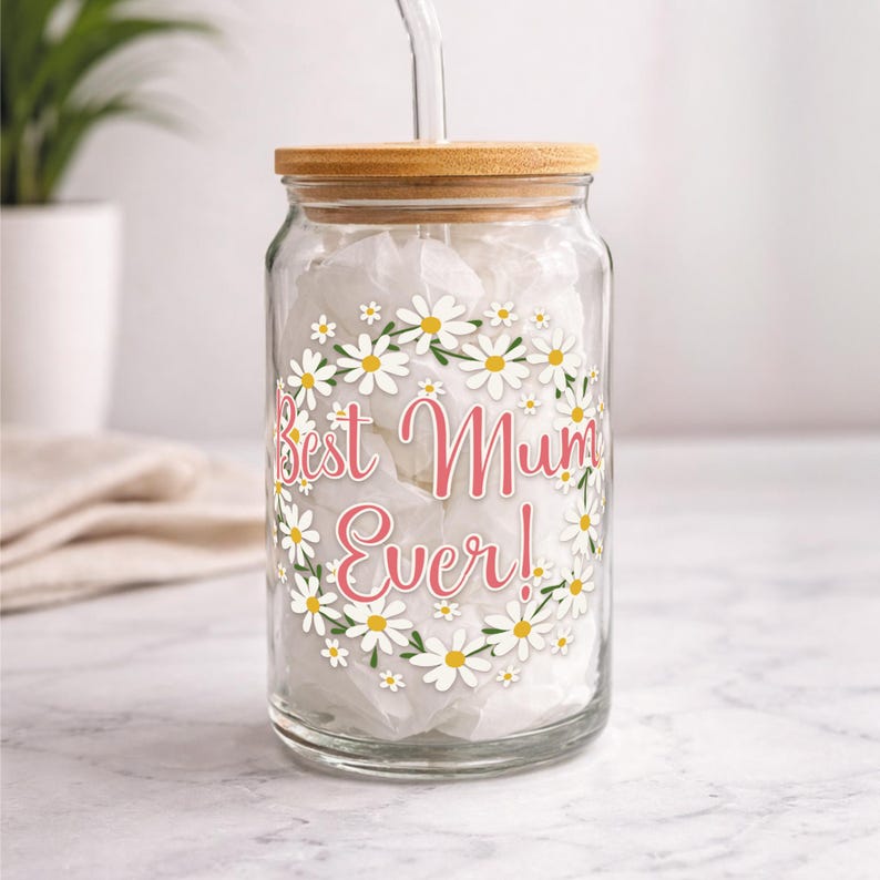 Puede incluir: Un tarro de cristal transparente con tapa de bamb&uacute; y pajita de cristal. El tarro est&aacute; decorado con una corona de margaritas y el texto "Best Mum Ever!" en rosa. El tarro est&aacute; relleno de papel de seda blanco.