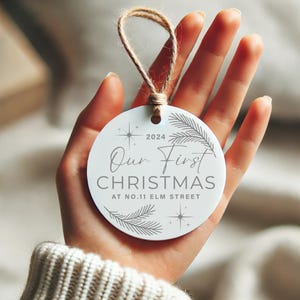 Op de afbeelding: Ronde witte kerstbal met de tekst "Our First Christmas at No. 11 Elm Street" en het jaar 2024. De kerstbal is versierd met een gestileerde dennentak en sterren.