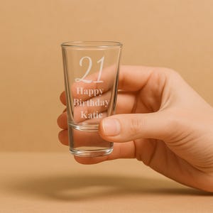 May include: A clear shot glass with the number "21" and the words "Happy Birthday Katie" etched on the front. The glass is held in a hand against a neutral background. The shot glass is cylindrical and appears empty.