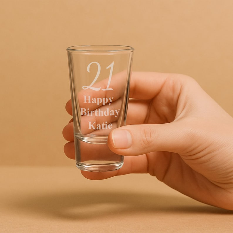 May include: A clear shot glass with the number "21" and the words "Happy Birthday Katie" etched on the front. The glass is held in a hand against a neutral background. The shot glass is cylindrical and appears empty.