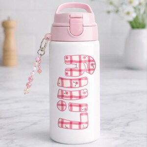Puede incluir: Una botella de agua blanca con tapa y asa rosas. La botella presenta la palabra "JUNE" en un patr&oacute;n de cuadros rosas y blancos con peque&ntilde;os detalles florales. Un dije de cuentas con cuentas rosas y transparentes cuelga de la botella.
