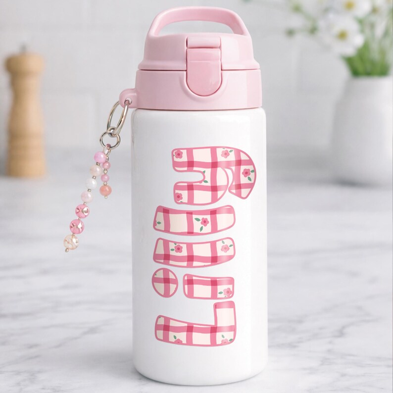 Puede incluir: Una botella de agua blanca con tapa y asa rosas. La botella presenta la palabra "JUNE" en un patr&oacute;n de cuadros rosas y blancos con peque&ntilde;os detalles florales. Un dije de cuentas con cuentas rosas y transparentes cuelga de la botella.