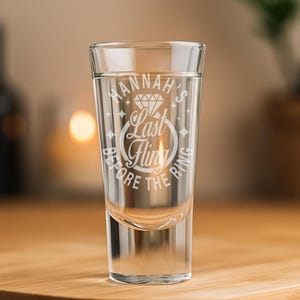 May include: A clear shot glass with the text "Hannah's Last Fling Before The Ring" etched in white. The glass is filled with a clear liquid and sits on a wooden surface. The background is blurred, with a candle and greenery visible.