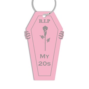 May include: Pink coffin-shaped keyring with a grey skeleton hand on each side. The keyring has a grey flower and the text "R.I.P. MY 20s" on it.