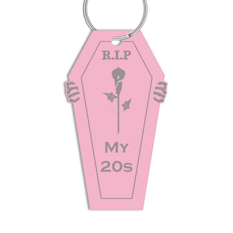 May include: Pink coffin-shaped keyring with a grey skeleton hand on each side. The keyring has a grey flower and the text "R.I.P. MY 20s" on it.