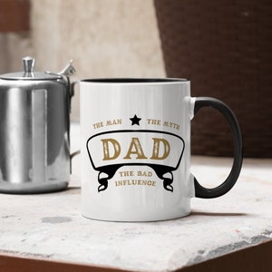 May include: White ceramic mug with a black rim and handle. The mug has a gold design that says "The Man, The Myth, Dad, The Bad Influence".