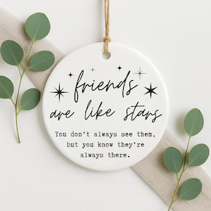 May include: A white ceramic ornament with the text "friends are like stars" in black script. The ornament has a jute string for hanging and is decorated with star graphics. Green eucalyptus sprigs and a beige cloth are in the background.