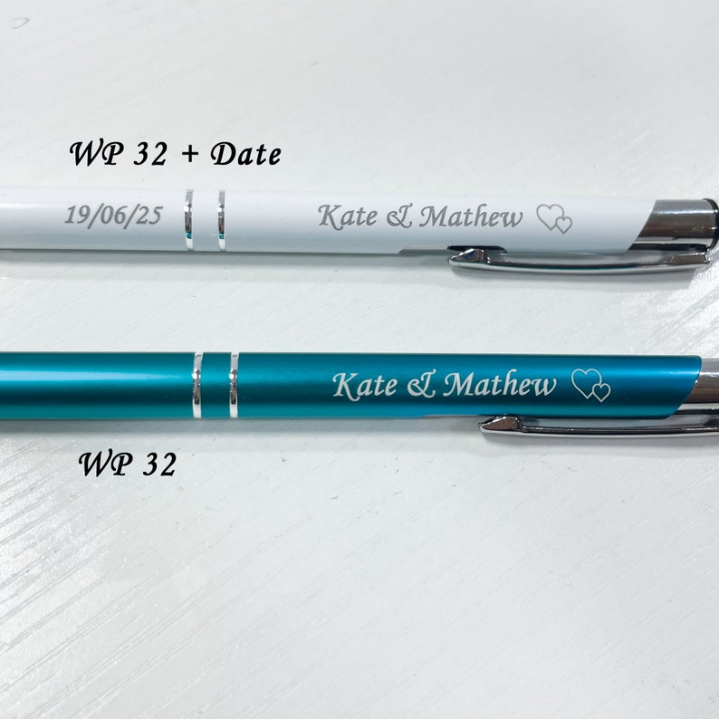 May include: Two pens with personalized engravings. The top pen is white with silver accents and the bottom pen is teal blue with silver accents. Both pens have the text "Kate & Mathew" and two hearts engraved on them. The top pen also has the text "WP 32 + Date" and "19/06/25" engraved on it. The bottom pen has the text "WP 32" engraved on it.