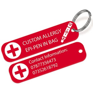 Op de afbeelding: Rode allergiewaarschuwingssleutelhanger met witte tekst: "CUSTOM ALLERGY EPI-PEN IN BAG." Bevat een wit kruissymbool en contactgegevens. Bevestigd aan een zilveren sleutelring.