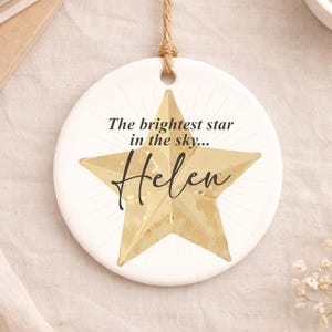 May include: A white, round ceramic ornament with a gold star design. The text "The brightest star in the sky... Helen" is printed on the ornament. A natural twine loop is attached for hanging.