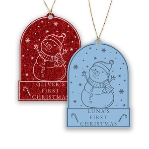 May include: Two Christmas ornaments featuring a snowman in a snow globe. The ornament on the left is red with glitter and says "Oliver's First Christmas". The ornament on the right is blue and says "Luna's First Christmas".