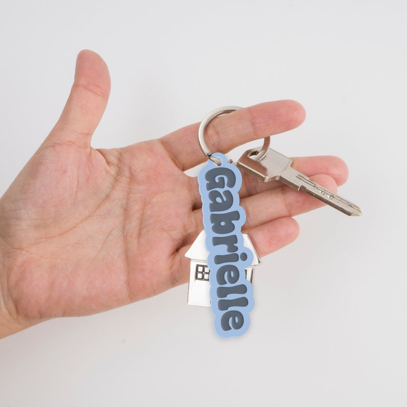 May include: A blue house-shaped keychain with the name "Gabrielle" printed in black letters. The keychain is attached to a silver key.