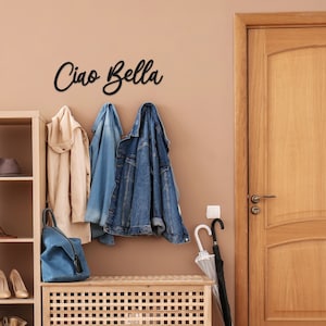 May include: A wooden bench with a slatted design sits in front of a beige wall with a black metal sign that reads "Ciao Bella".  Three jackets hang on hooks on the wall, one beige, one blue denim, and one dark blue denim.  A blue backpack sits on the bench.