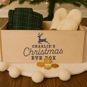 Pu&ograve; includere: Una scatola di legno per la vigilia di Natale con la scritta "Charlie's Christmas Eve Box" e una grafica di renna. La scatola contiene un pigiama a quadri verdi e guanti color crema. Monete d'oro sono sparse davanti alla scatola.