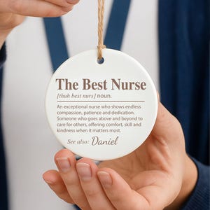 May include: A white, round ceramic ornament with the text "The Best Nurse" in a serif font. The ornament includes a definition of a nurse and the name "Daniel". A brown string is attached for hanging.