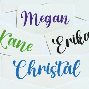 May include: Four white cards with names written in cursive on them. The names are Megan in purple, Kane in green, Erika in black, and Christal in blue.