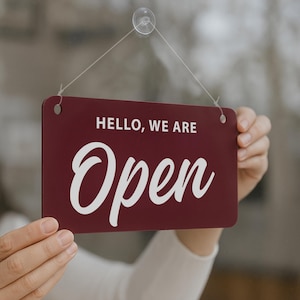 Op de afbeelding: Een bordeauxrode rechthoekige bewegwijzering met de woorden "HELLO, WE ARE Open" in witte letters. Het bord wordt opgehangen door een transparante zuignap en een touwtje. Het bord wordt door een persoon vastgehouden.
