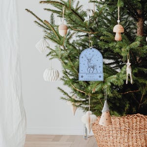 May include: Blue and white Christmas ornament with a reindeer and the text "Kelsey's First Christmas". The ornament is hanging on a Christmas tree with other ornaments.