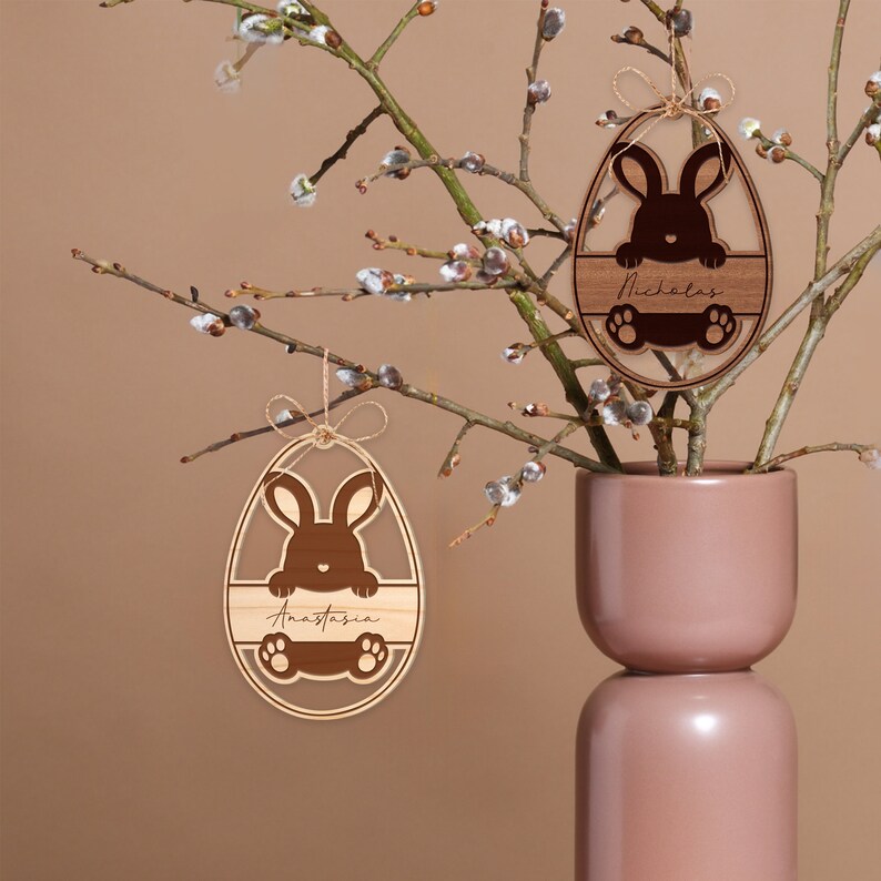 May include: Two wooden Easter egg ornaments with bunny cutouts and personalized names. The ornaments are hanging from a branch with white blossoms. The names on the ornaments are Anastasia and Nicholas.