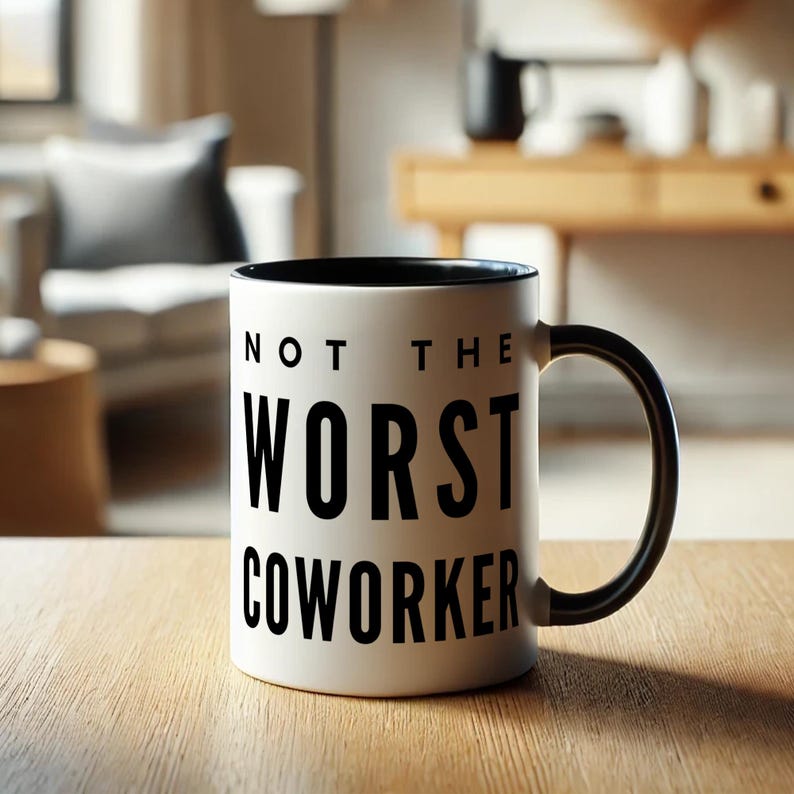 May include: White ceramic coffee mug with a black rim and handle. The mug has black text that reads "Not the Worst Coworker".