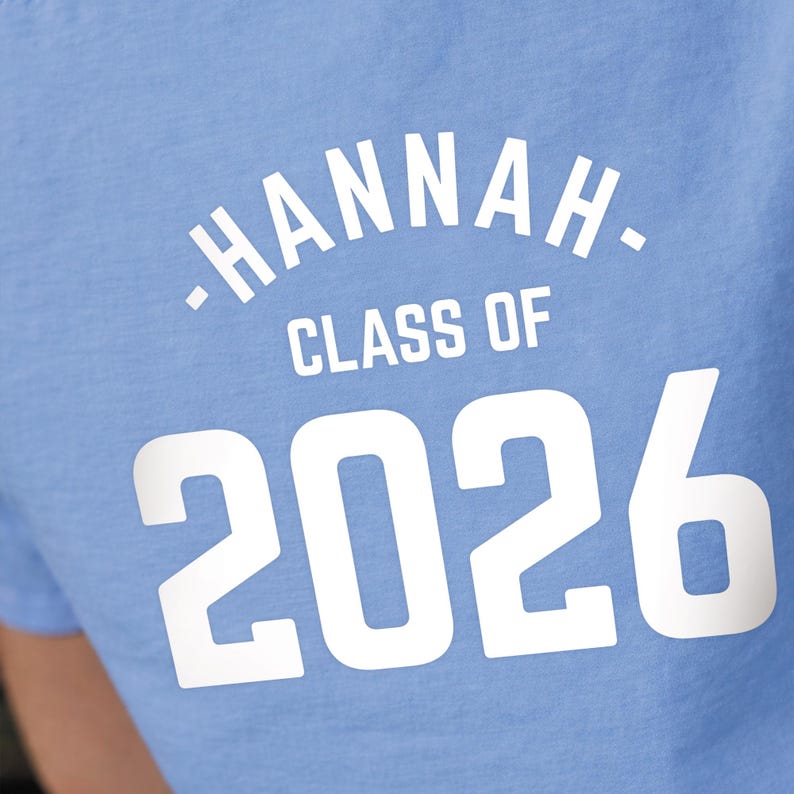 May include: Light blue t-shirt with the name "Hannah" arched across the top in white, followed by "Class of" and the year "2026" in a bold, white font. The shirt is made of a soft material.