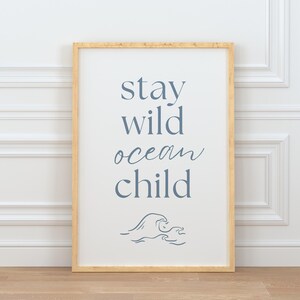 K&ouml;nnte beinhalten: Ein gerahmter Druck mit dem Text "stay wild ocean child" in blauer Schrift. Unter dem Text befindet sich eine stilisierte Wellengrafik.