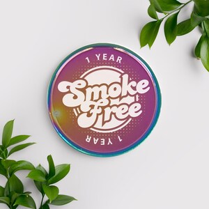 May include: A round, iridescent purple, pink, and blue sticker with the text "1 Year Smoke Free" in white. The sticker has a dotted pattern around the edge.
