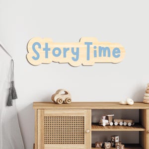 May include: A wooden sign reading "Story Time" in light blue, cloud-shaped letters. The sign is mounted on a white wall. A wooden toy car and a cabinet are positioned below the sign, adding to the nursery theme.