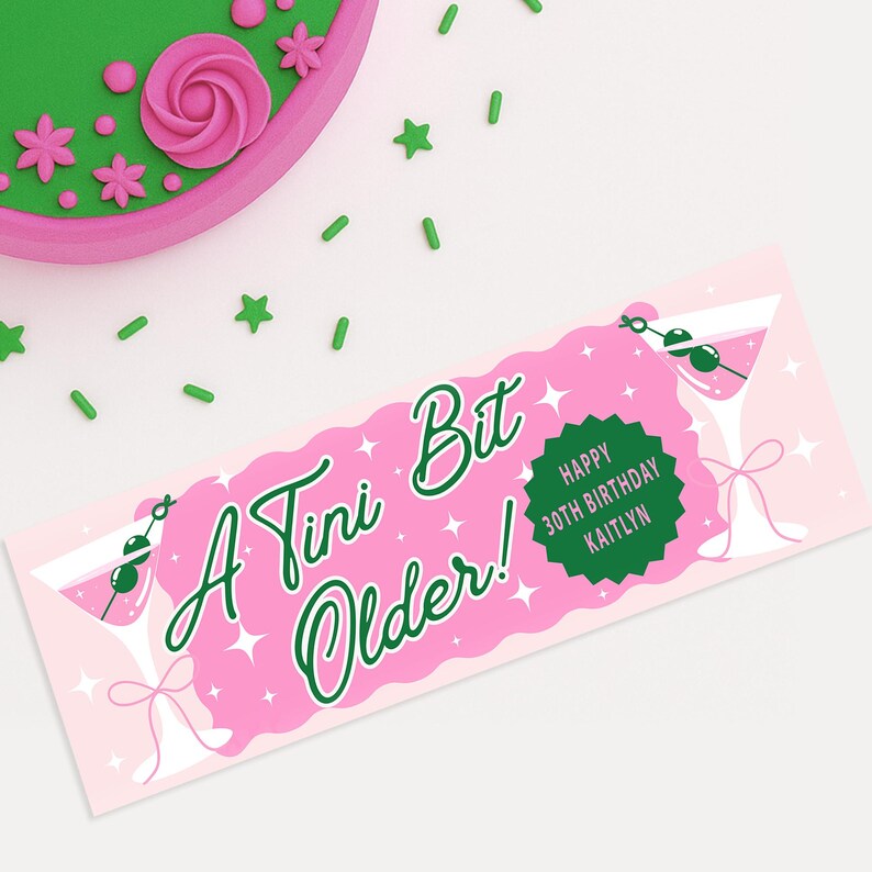 Op de afbeelding: Een roze banner met de tekst "A Tini Bit Older!" in groen schrift, martiniglazen en de woorden "Happy 30th Birthday Kaitlyn" in een groene cirkel. Groene stervormige sprinkles en een roze en groene taart zijn ook zichtbaar.