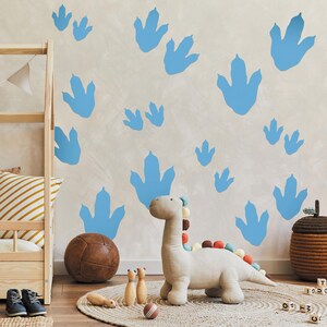 May include: A light blue dinosaur footprint wall decal set with a stuffed dinosaur toy in a nursery setting. The decals are on a light gray wall with a wooden shelf and a brown ball on the floor.
