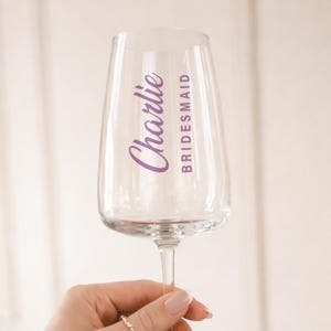 May include: A clear champagne flute with the name "Charlie" in a purple script font and the word "Bridesmaid" in a purple sans-serif font. The glass is held up against a soft, neutral background.