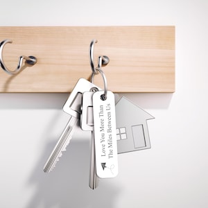 May include: Silver key chain with two keys and a house-shaped charm. The charm has the text "Love You More Than The Miles Between Us" on it.