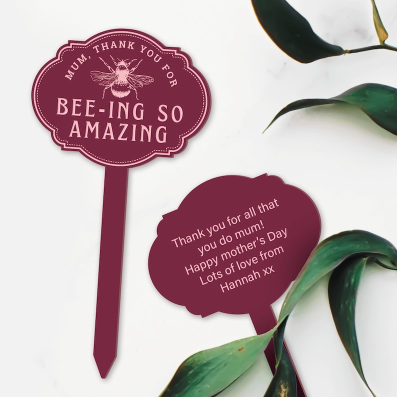 May include: Two burgundy garden stakes with pink text. The first stake says "Mum, Thank You For Bee-ing So Amazing" with a bee illustration. The second stake says "Thank you for all that you do mum! Happy Mother's Day Lots of love from Hannah xx".