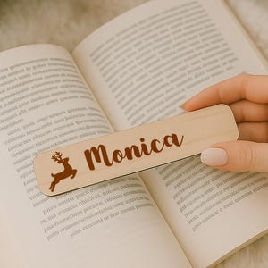 May include: A wooden bookmark with the name "Monica" in a brown script font and a brown reindeer silhouette. The bookmark is held over an open book with text.