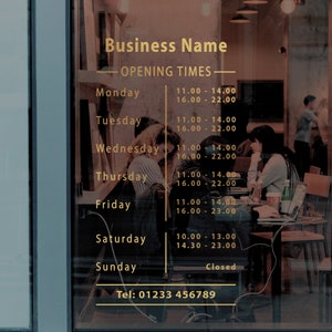 May include: A gold sign with the text "Business Name" and "Opening Times" with the hours of operation listed for each day of the week. The sign is on a glass door with a blurry image of people sitting at tables in the background.